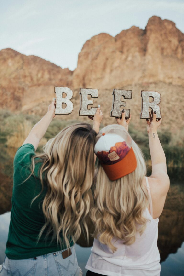 Two women holding 'BEER' letters with a scenic outdoor backdrop, conveying a fun and relaxed vibe.
