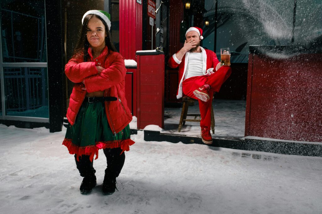 Santa and a festive elf in winter attire outside a red building, embodying holiday cheer.