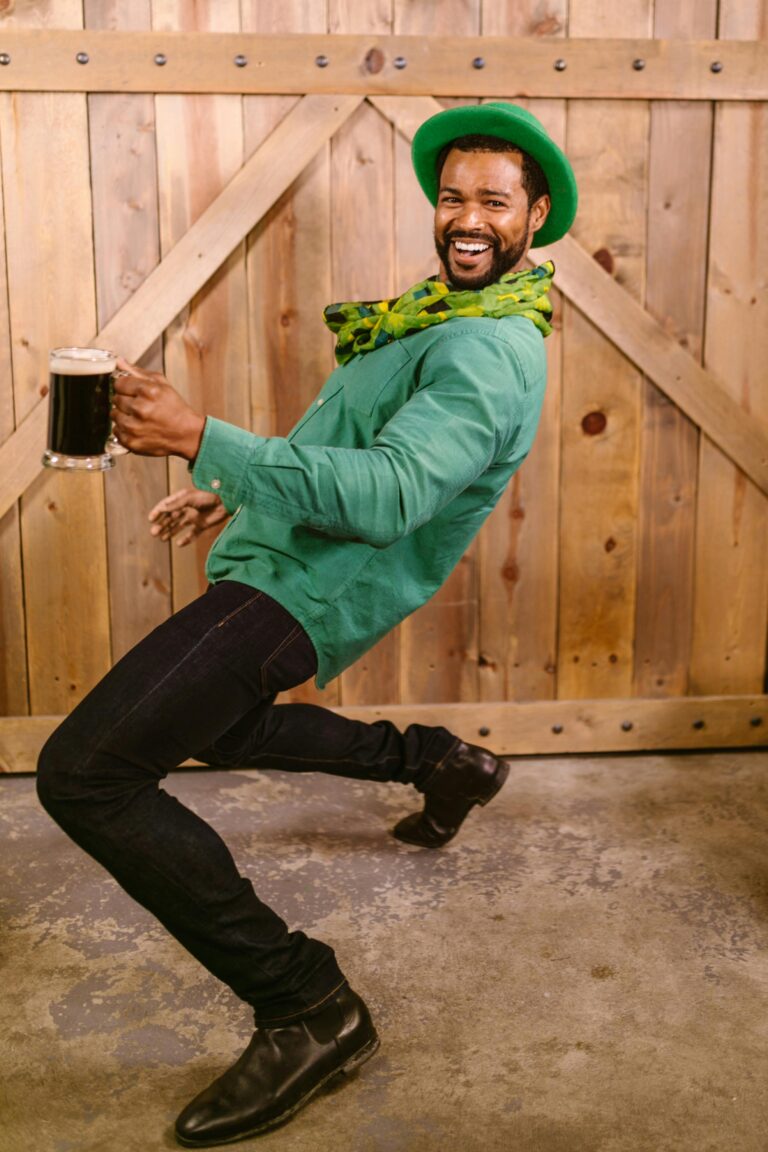 Smiling man in green outfit with beer, posing playfully indoors for St. Patrick's Day.